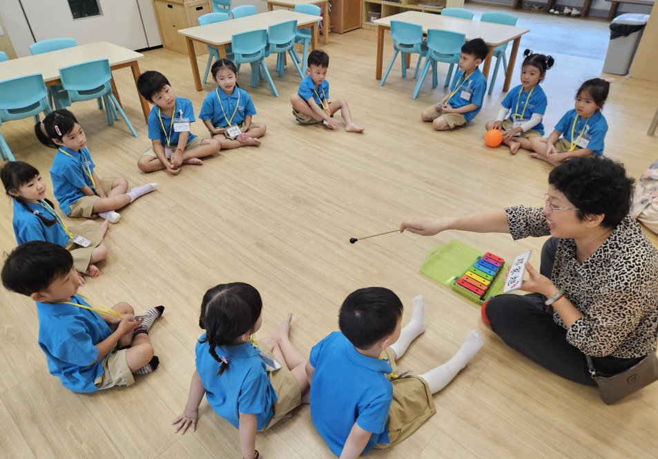 Teacher holds a mallet by a xylophone as young children in blue uniforms sit in a circle on a classroom floor.